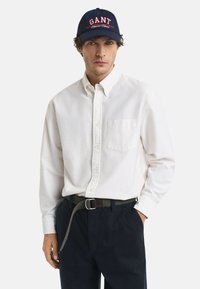 Camicia bianca a maniche lunghe con taschino sul petto, abbinata a pantaloni scuri e un cappellino blu navy con lettere rosse. Design e texture semplici.