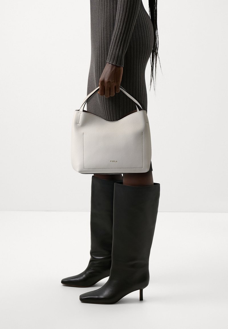 Vit läderhandväska med en slät yta, rundad topp och en framficka. Handtagen är diskreta. Modellen bär en grå rippad klänning och höga svarta stövlar.