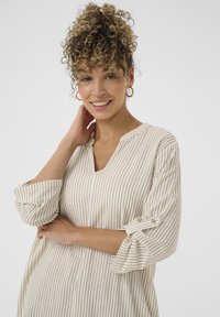 Blusa beige a righe con scollo a V, maniche arrotolabili e texture morbida. Presenta sottili linee verticali e un design con vestibilità rilassata.