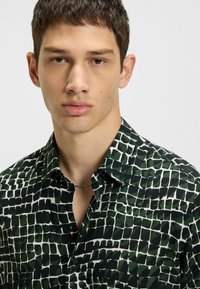 Camicia con bottoni caratterizzata da un motivo astratto verde e nero su un tessuto liscio, con colletto a punta e un sottile design della scollatura aperta.