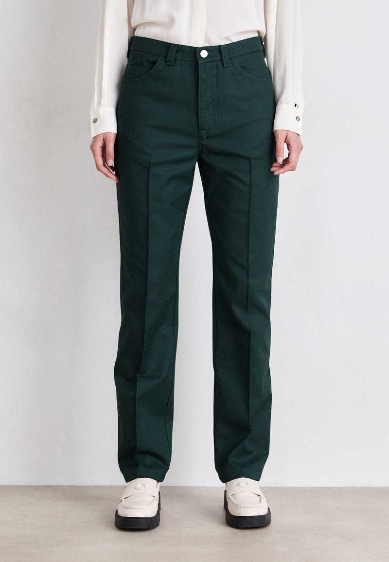 Personne portant un pantalon vert foncé sur mesure avec des plis sur le devant, une chemise blanche boutonnée et des mocassins blancs, debout sur un sol clair contre un mur uni.