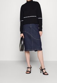 Žena v černém topu s dlouhým rukávem a logem, tmavé džíny midi sukně, černých sandálech na vysokém podpatku a s černou kabelkou.