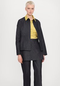 Giacca di jeans scura con zip frontale, abbinata a una camicia gialla con bottoni e pantaloni scuri coordinati. Linee pulite e design semplice.