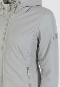 Giacca grigia con cappuccio realizzata in un tessuto liscio e testurizzato. Include una cerniera frontale, tasche laterali e un dettaglio del logo sulla manica.