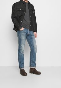 Schwarze Denim-Hemd mit zwei Brusttaschen, graues T-Shirt darunter, hellblaue, ausgewaschene Jeans und braune Leder-Stiefeletten.