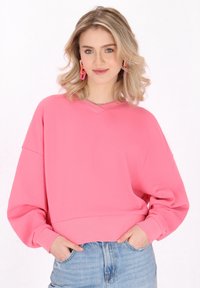 Sudadera rosa corta con escote en V, hombros caídos y puños acanalados. Se lleva con jeans de mezclilla clara de tiro alto. El fondo es blanco liso.