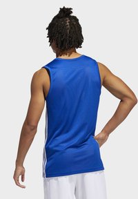 Manlig idrottare med mörkt lockigt hår bär en blå ärmlös sporttröja och vita shorts, står med en hand på höften, ryggen vänd mot betraktaren.