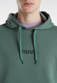 Felpa verde realizzata in un mix di cotone con un tessuto testurizzato. Presenta un logo ricamato e dei lacci, con una maglietta viola a strati sotto.