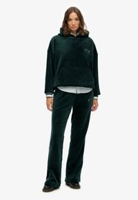 Hoodie en velours vert avec grande poche avant et capuche avec cordon de serrage, associé à un pantalon en velours assorti. Les deux présentent des détails de logo discrets.