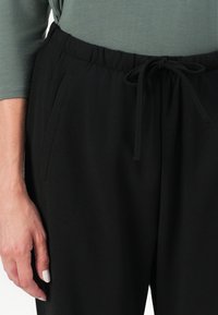 Sorte joggebukser med elastisk midje og snor, med sidelommer og en glatt tekstur. Nærbilde med en hånd.