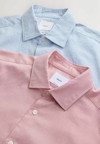 Deux chemises à col pliées, une bleu clair à rayures blanches, une rose uni, toutes deux avec des boutons blancs et des étiquettes de la marque « NEXT ».