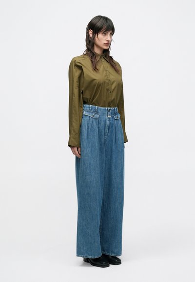 Πουκάμισο σε ελιά πράσινο με κουμπιά και μακριά μανίκια, συνδυασμένο με φαρδιά παντελόνια τζιν σε ανοιχτό γαλάζιο και μαύρα παπούτσια. Λείο ύφασμα και άνετη εφαρμογή.