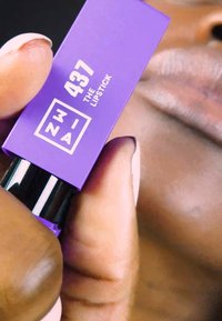 Tube de rouge à lèvres violet étiqueté "437 THE LIPSTICK", présentant un design épuré avec des accents noirs, tenu près des lèvres d'une teinte neutre.
