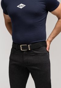 Mężczyzna w granatowej koszulce Lee Cooper schowanej w ciemnoszare spodnie z czarnym skórzanym paskiem, stojący z jedną ręką w kieszeni.