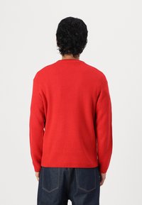 Pull en tricot rouge avec une finition texturée, col rond et manches longues, associé à un pantalon en denim foncé.