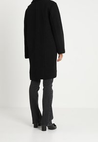 Černý huňatý kabát s rovnými liniemi, vysokým límcem a volným střihem. Nosič má na sobě tmavé zvonové džíny a černé boty. Textura vypadá měkce.