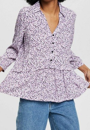 Femme portant un chemisier ample violet à motifs floraux avec des boutons, associé à un jean en denim bleu, tenant l'ourlet du chemisier des deux mains.