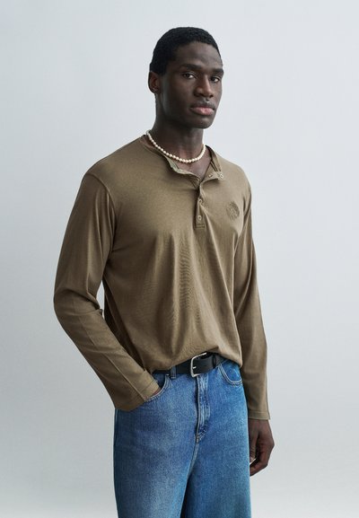 Mann trägt braunes Langarmshirt mit Perlenkette und blauer Jeans, steht mit einer Hand in der Tasche vor einfachem grauem Hintergrund.