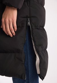 Czarna kurtka puchowa z bocznym zamkiem, wyraźnym wzorem pikowania i gładką podszewką. Ręka spoczywa blisko zamka, gotowa do interakcji.