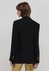 Blazer nero con una vestibilità su misura, spacco posteriore singolo e dettagli in metallo sui polsini. Tessuto morbido e spalle strutturate.