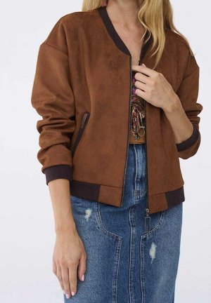 Donna che indossa una giacca marrone con zip e bordi scuri sopra una maglia fantasia e una gonna in denim blu consumata.
