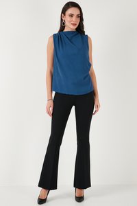 Kvinde med langt mørkt hår iført ærmeløs blå draperet top, sorte tætsiddende flarebukser, sorte hæle og sølvfarvede hoop-øreringe mod hvid baggrund.