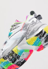Białe buty sportowe z multikolorową podeszwą o zygzakowatych wzorach, teksturowaną cholewą z neonowymi akcentami oraz widocznym logo z boku.