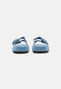 Blå jeanssandaler med platt sula, öppen baksida och fransade tygdetaljer. Har en justerbar rem på varje sida.