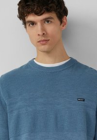 Pull bleu texturé avec un col rond, des motifs rayés et une petite étiquette logo sur la poitrine. Tissu doux avec un design décontracté.