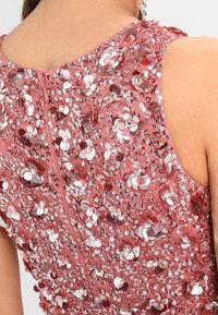 Šaty z tkaniny s flitry v podobě růže, s kulatým výstřihem a zapínáním na zip na zadní straně. Textura vykazuje lesklé stříbrné a růžové kruhy po celé ploše.