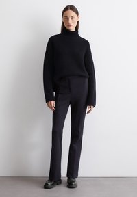 Schwarzer, gerippter Rollkragenpullover mit langen Ärmeln, kombiniert mit eng geschnittenen schwarzen Hosen und schwarzen, klobigen Lederstiefeln, vor einem neutralen Hintergrund.