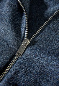 Närbild av en delvis öppnad metalldragkedja på mörkblå, strukturerad tyg, som visar dragkedjans tänder och dragtabbens detaljer.