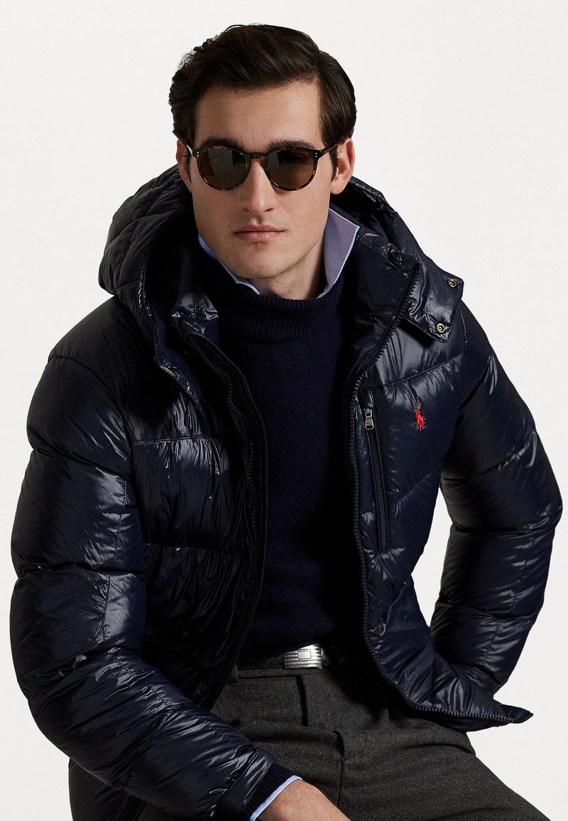 Marine blauwe pufferjack met een capuchon, glanzend materiaal, ritssluiting, klein logo op de borst, gedragen over een marine blauwe trui, gecombineerd met donkere zonnebril.