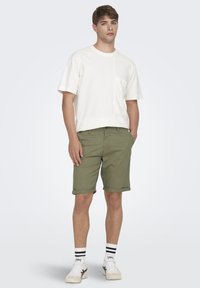 T-shirt blanche à manches courtes avec une poche, associée à un short olive avec des ourlets roulés, et des baskets blanches avec des rayures noires sur les chaussettes.