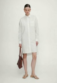 Valge puuvillane kleit nööbiga, pikkade varrukatega ja rinna taskuga. Mudel hoiab käes punutud pruuni kotti ja kannab beeže sandaale.