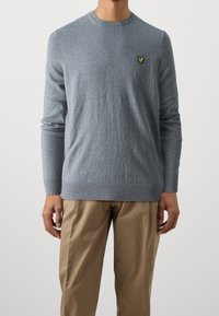 Homme portant un pull gris ajusté à manches longues avec un logo d'oiseau jaune sur la poitrine et un pantalon beige sur un fond uni.