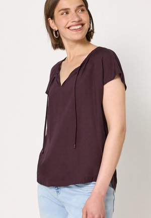 Jeune femme souriante, portant un chemisier violet foncé à manches courtes avec un col en V et un jean bleu clair sur un fond clair uni.