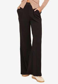 Zwarte wijde broek met geplooide voorkant en zijzakken, gemaakt van een gladde stof, gecombineerd met een beige cardigan en casual schoenen.