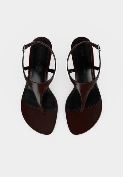 Para ciemnobrązowych skórzanych sandałów typu thong z paskami wokół kostki i małymi klamerkami, płaska podeszwa, widoczna z góry na białym tle.