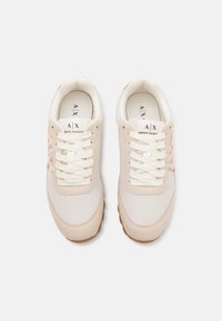 Baskets de sport blanches et beiges avec des panneaux en maille, des semelles en caoutchouc texturé et des lacets crème. Présente un détail de logo rose pâle sur les côtés.