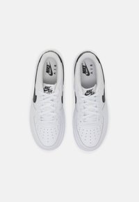 Paire de baskets blanches Nike Air Force 1 avec des logos swoosh noirs, vues de dessus, montrant les lacets et les zones perforées à l'avant.