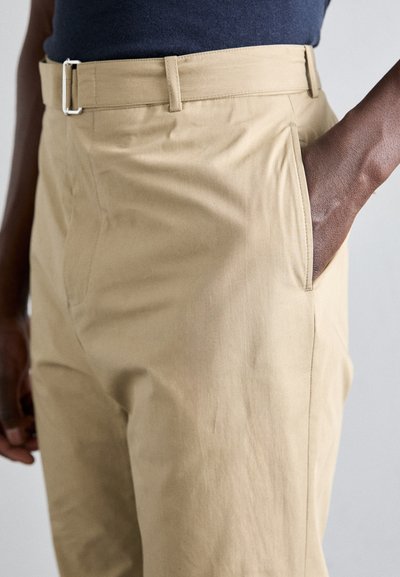 Pantalons beige clair dotés d'une texture lisse, d'un devant plat, de poches latérales et d'une boucle en métal argenté sur la ceinture.