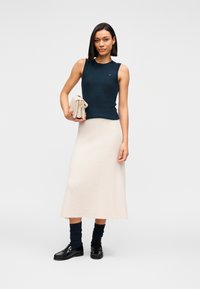 Ναυτικό μπλε αμάνικο τοπ, υφασμάτινη μπεζ μίντι φούστα, μαύρα μπαλαρίνες και ναυτικό μπλε κάλτσες. Το μοντέλο κρατάει ένα ανοιχτό μπεζ τσαντάκι. Απλός, μοντέρνος σχεδιασμός.