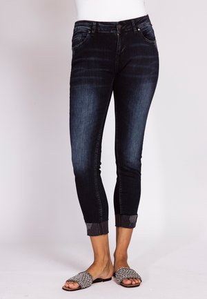 Frau trägt dunkelblaue Skinny-Jeans mit aufgerollten Bündchen und schwarz-weiße, gewebte Slip-on-Sandalen, steht vor einem einfarbigen Hintergrund.