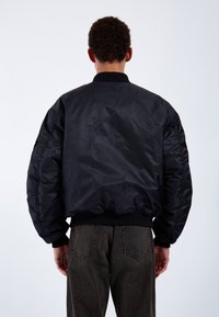 Black bomber jacket with a smooth, shiny exterior, ribbed cuffs and hem, and oversized, puffy sleeves. Worn over dark denim pants.
