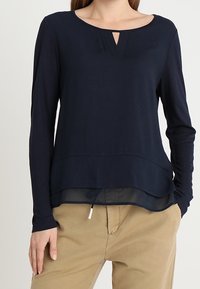 Femme portant un haut bleu marine à manches longues avec un détail en tissu transparent à l'ourlet et un pantalon beige, debout avec une main dans la poche devant un fond uni.