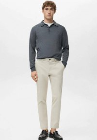 Młody mężczyzna stojący, ubrany w ciemnoszarą koszulkę polo z długim rękawem, beżowe spodnie i czarne mokasyny na jednolitym białym tle.