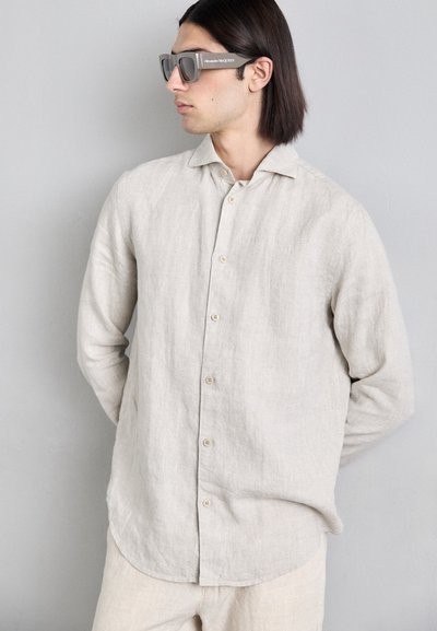 Chemise en lin beige avec un devant boutonné, coupe décontractée et ourlet arrondi. Le modèle porte des lunettes de soleil grises surdimensionnées.
