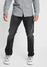 Vaqueros de mezclilla gris oscuro con un ajuste ceñido, con un sutil desgaste. Combinados con zapatillas blancas y una camisa gris claro abotonada.