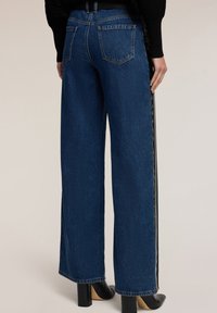 Szerokie, ciemnoniebieskie dżinsy z czarnymi paskami po bokach i kieszeniami z tyłu. Dopasowany stan i standardowe szwy na brzegach. Widoczne słupkowate obcasy.